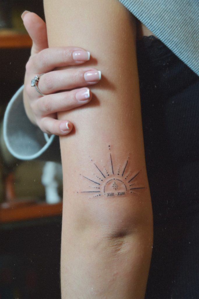 Un bras avec un tatouage représentant un soleil et une date stylisé.